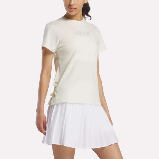 A woman stands against a plain light background wearing the Reebok Women's Ribbon T-Shirt in Linen, a short-sleeved cotton top featuring small bow details on the side, paired with a white pleated skirt.