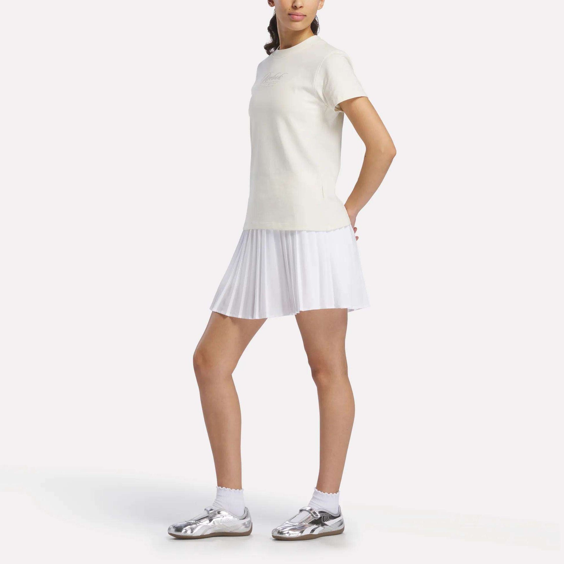 A person wearing a Reebok Women's Ribbon T-Shirt paired with a white pleated skirt, white socks, and shiny silver shoes stands against a plain background.