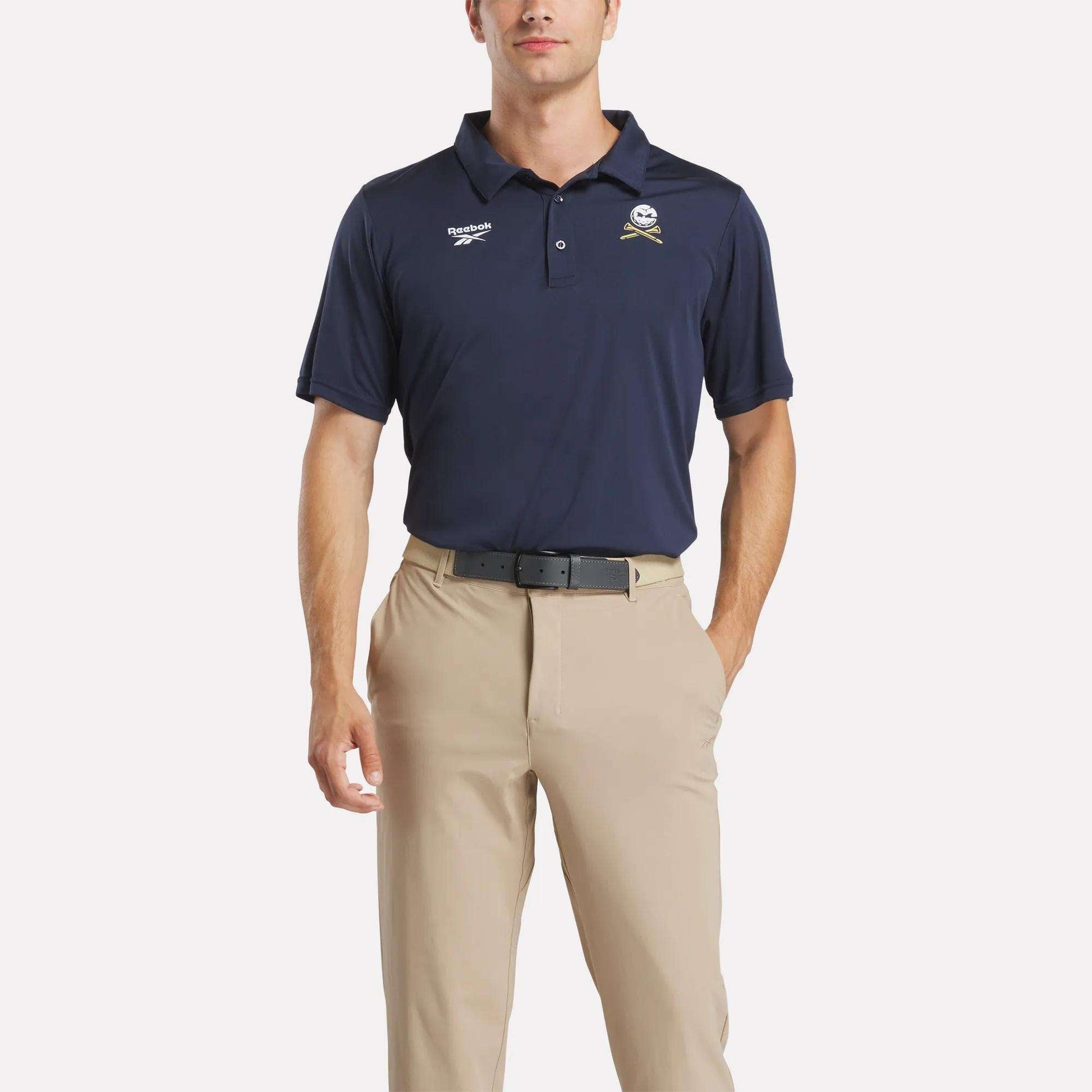 A man stands against a plain white background wearing the Reebok Men's Crushers GC Replica Polo in navy blue, featuring embroidered logos, paired with tan khaki pants and one hand in his pocket.