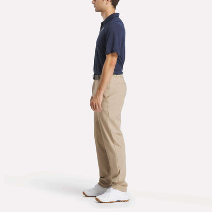 A man stands in profile against a white background, wearing a Reebok Men's Crushers GC Replica Polo in navy, beige khaki pants, a black belt, and white sneakers.