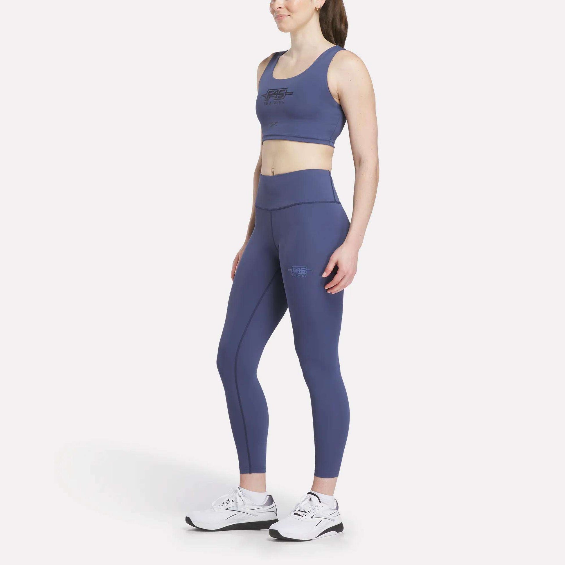 Outfitted in the Women's Reebok x F45 Training Sports Bra, a woman stands and smiles against a plain white background, paired with matching blue leggings and white athletic shoes—ready for her next F45 Training session.