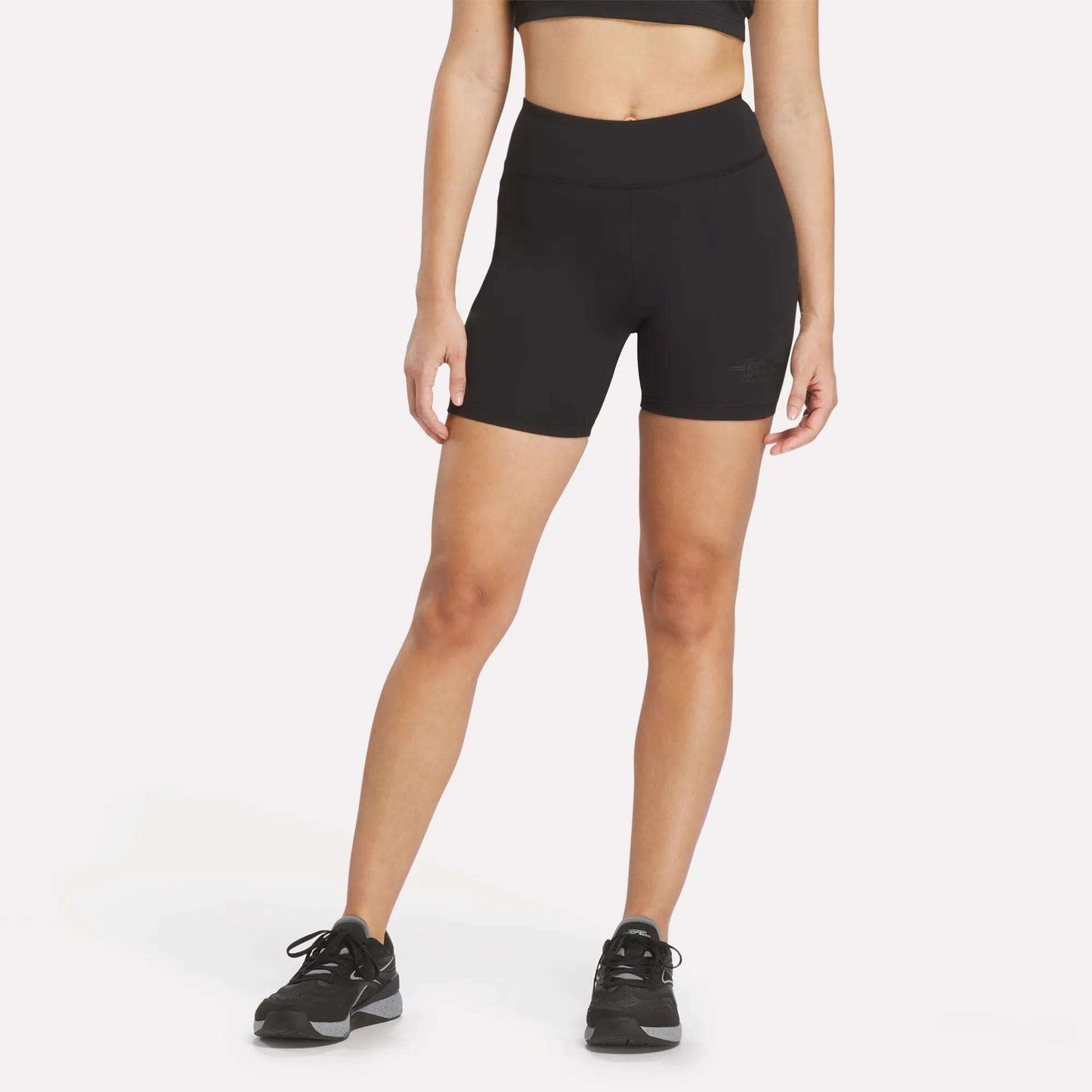 A woman wearing a black sports bra, Women's Reebok x F45 Training High-Rise Biker Shorts, and black running shoes stands against a plain white background.