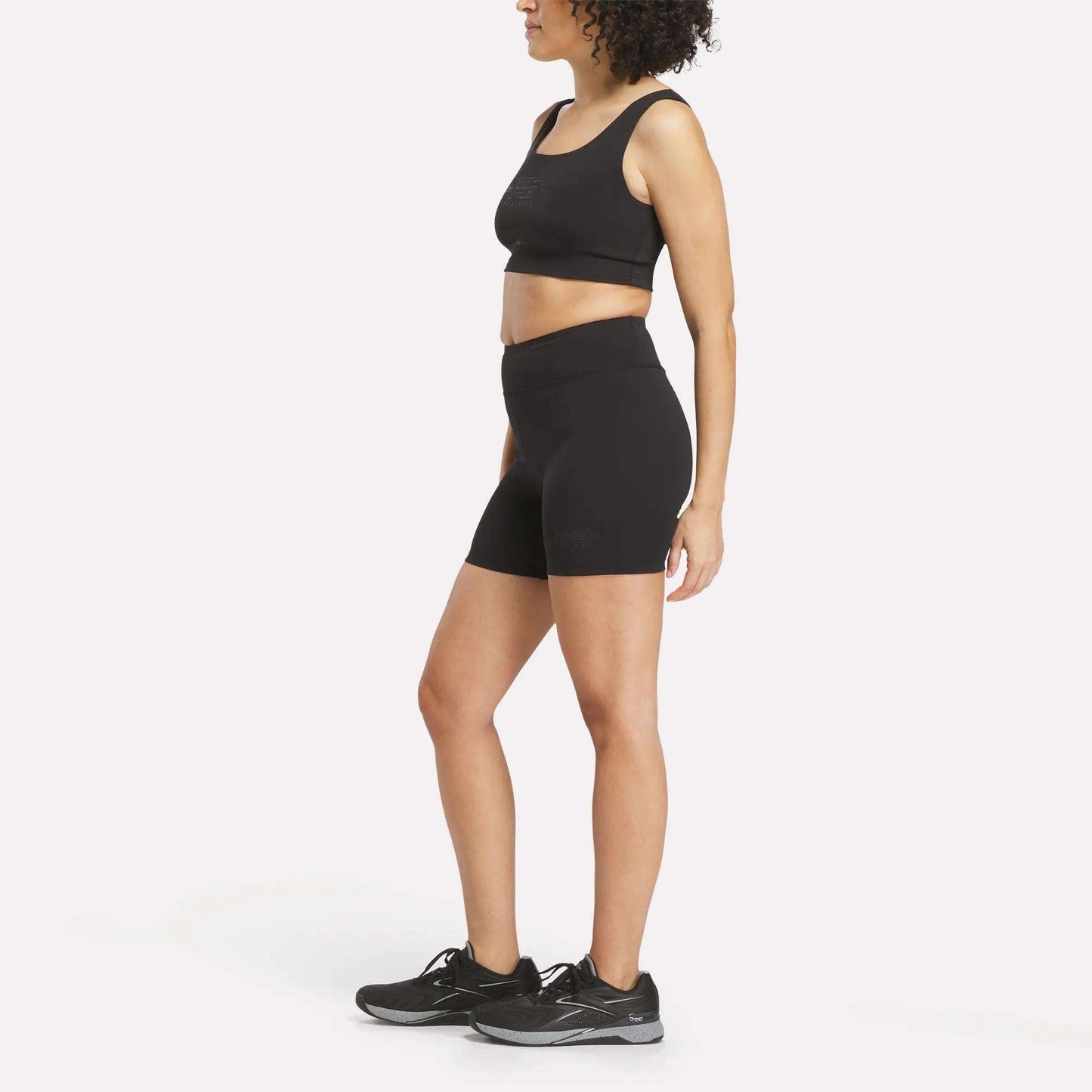A woman stands against a plain white background, facing sideways with her arms relaxed, wearing a black Reebok performance sports bra, Women's Reebok x F45 Training High-Rise Biker Shorts in black, and black running shoes.