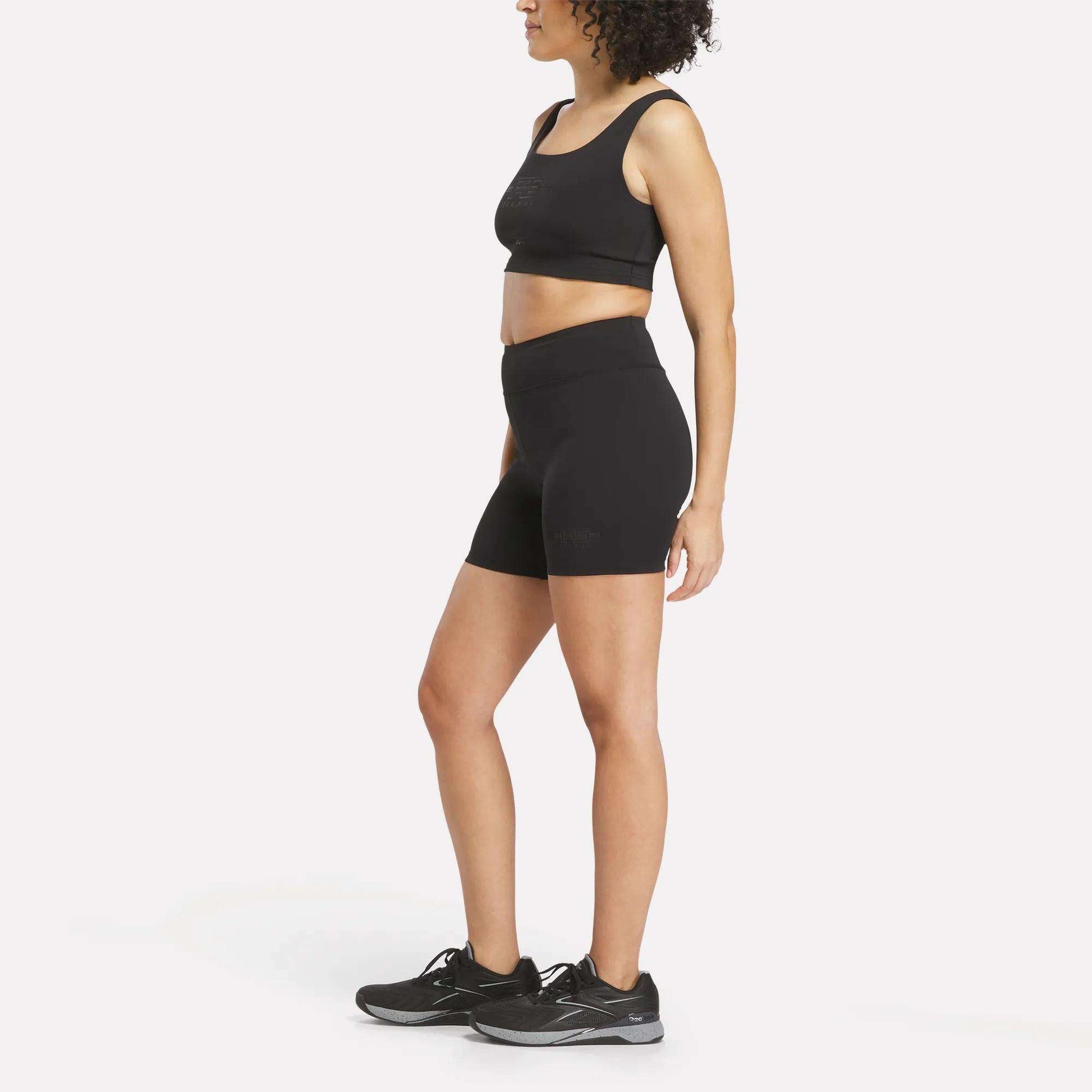 A woman stands against a plain white background, facing sideways with her arms relaxed, wearing a black Reebok performance sports bra, Women's Reebok x F45 Training High-Rise Biker Shorts in black, and black running shoes.