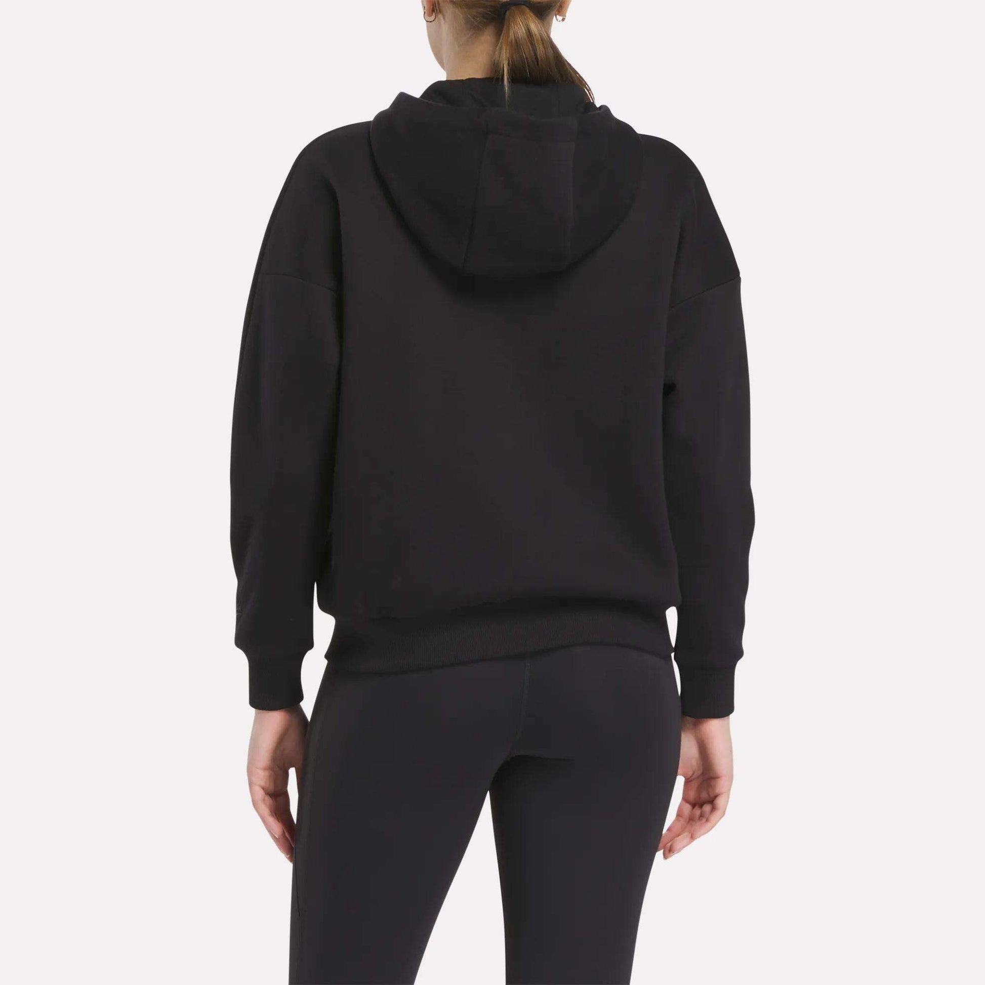 A person is shown from the back wearing a black Relaxed Reebok x F45 Women's Zip Hoodie by Reebok and black leggings. The hood is down, their hair is in a ponytail, and the background is plain light gray.