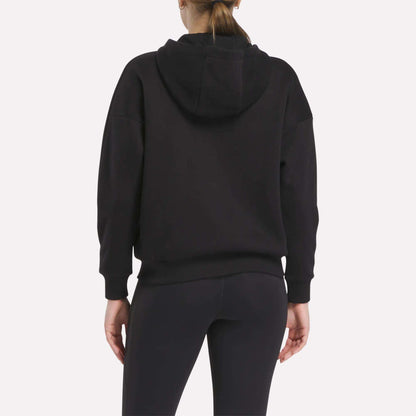 A person is shown from the back wearing a black Relaxed Reebok x F45 Women's Zip Hoodie by Reebok and black leggings. The hood is down, their hair is in a ponytail, and the background is plain light gray.