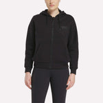 A person faces forward wearing a black Relaxed Reebok x F45 Women's Zip Hoodie by Reebok with front pockets and hood, paired with black pants. The background is plain and light-colored.