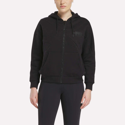A person faces forward wearing a black Relaxed Reebok x F45 Women's Zip Hoodie by Reebok with front pockets and hood, paired with black pants. The background is plain and light-colored.