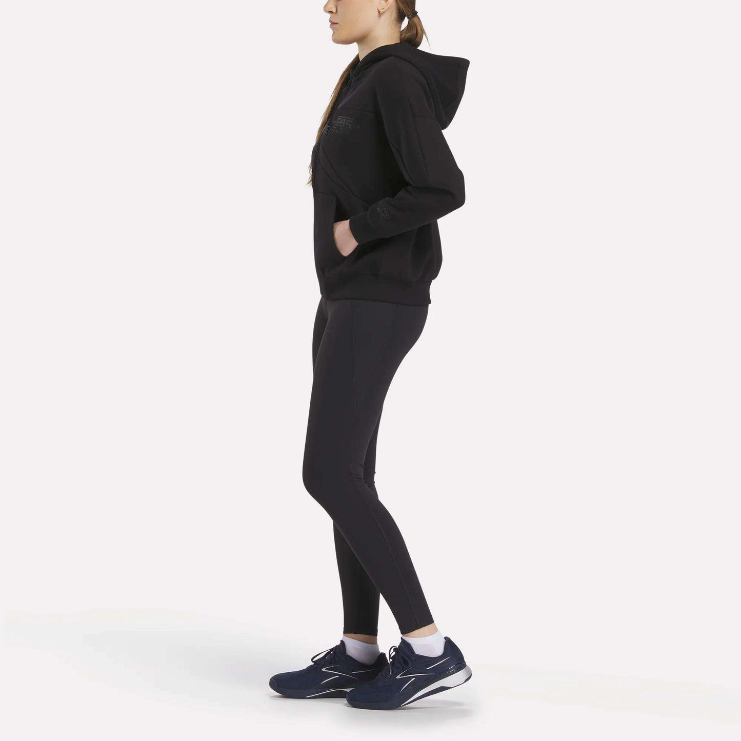 A woman wearing a Relaxed Reebok x F45 Women's Zip Hoodie stands in profile with her hands in her pockets, paired with black leggings and navy blue sneakers, against a plain white background.