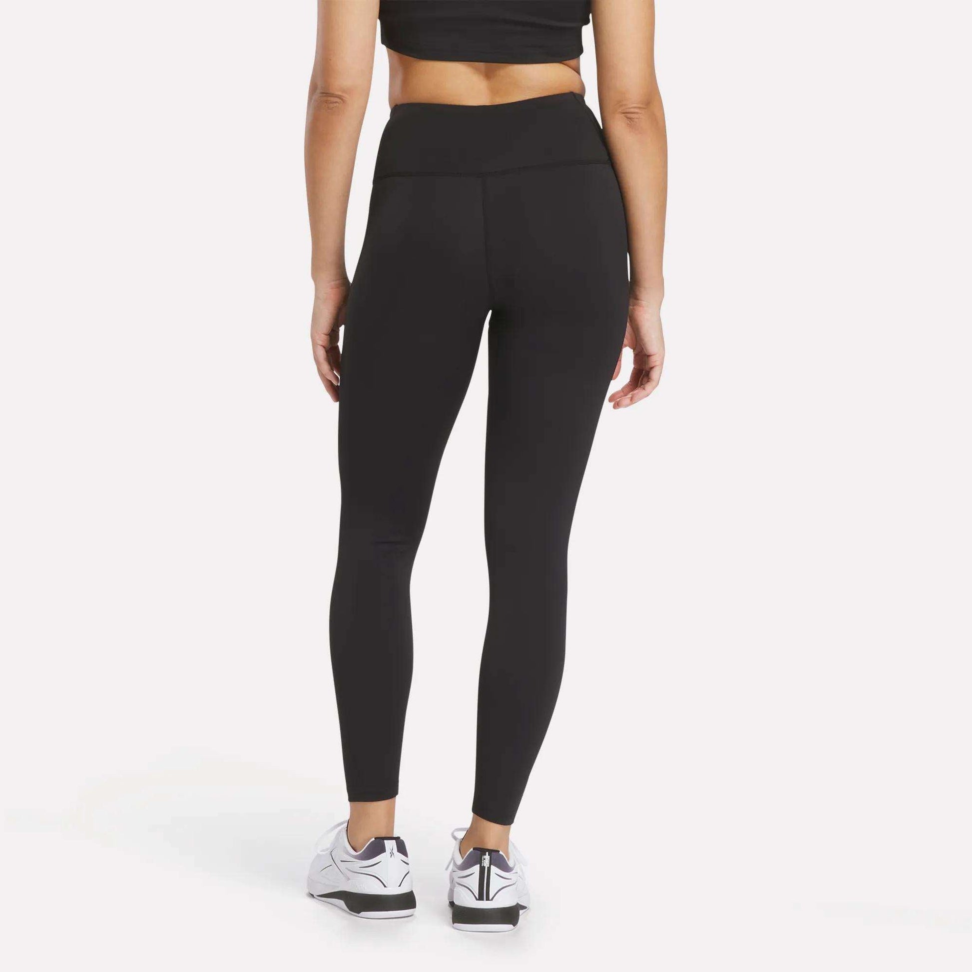 A person wearing a black crop top, Women's Reebok x F45 Training Leggings, and white Reebok performance athletic shoes stands facing away against a plain white background.