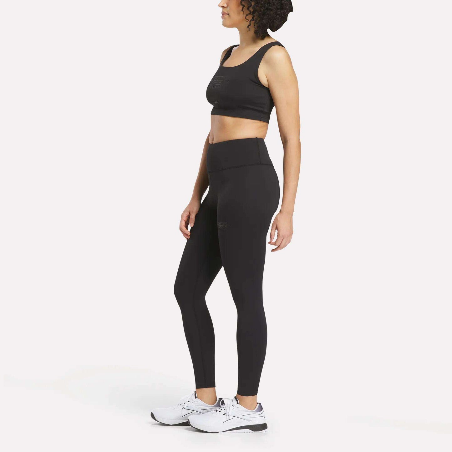 A woman stands in profile against a plain white background, wearing black high-waisted Women's Reebok x F45 Training Leggings and white Reebok performance sneakers.