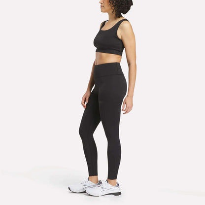 A woman stands in profile against a plain white background, wearing black high-waisted Women's Reebok x F45 Training Leggings and white Reebok performance sneakers.