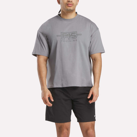 A man wearing a loose Reebok x F45 Training Graphic T-Shirt in gray and black shorts stands facing forward against a plain white background.