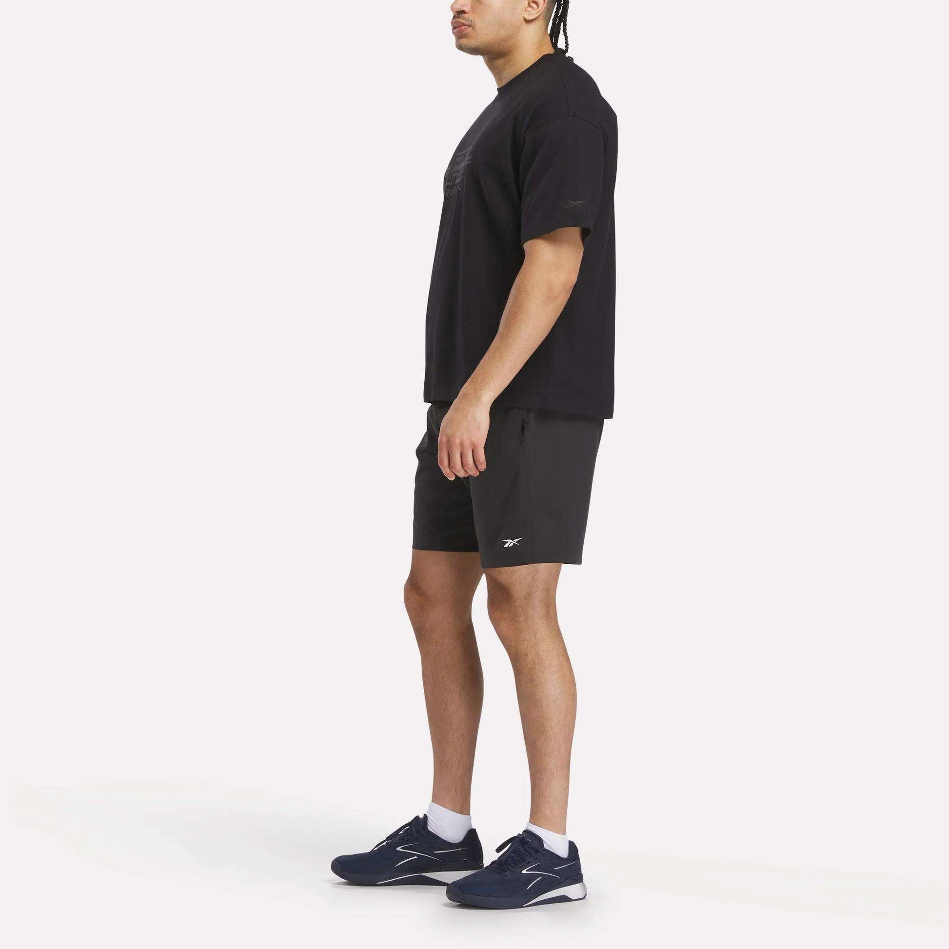 A man stands sideways against a plain white background wearing a Reebok x F45 Training Tonal Graphic T-Shirt, black athletic shorts, white socks, and dark blue sneakers.