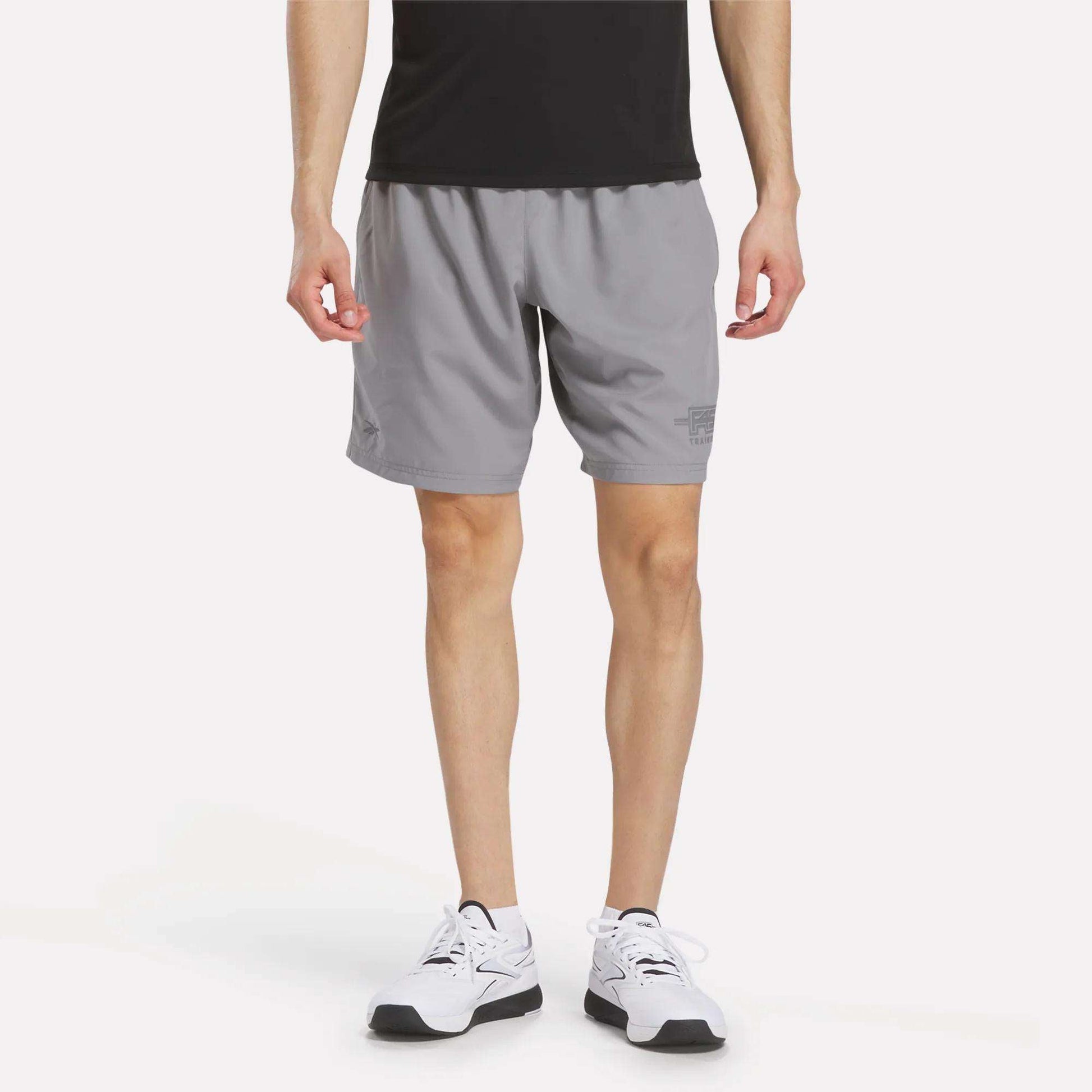 A person in a black shirt, Men's Reebok x F45 Training Woven Shorts (gray), and white running shoes stands against a plain white background, shown from shoulders to feet—ready for intense workouts or F45 Training.