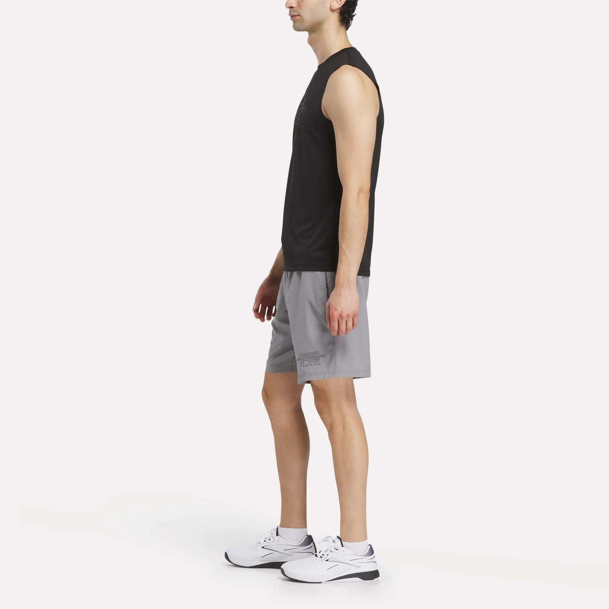 A man in a sleeveless black shirt, Reebok Men's Reebok x F45 Training Woven Shorts, and white sneakers stands in profile against a plain white background, ready for high-intensity training.