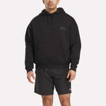 A man wears a Men's Reebok x F45 Training Relaxed Hoodie in black with a small chest logo and black athletic shorts, standing against a plain white background.