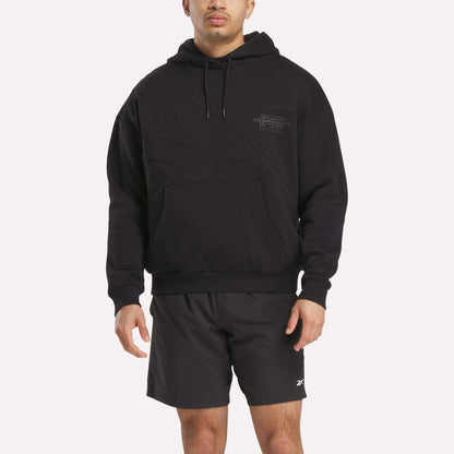 A man wears a Men's Reebok x F45 Training Relaxed Hoodie in black with a small chest logo and black athletic shorts, standing against a plain white background.