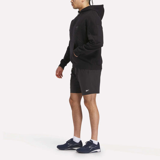 A person in profile wears the Men's Reebok x F45 Training Relaxed Hoodie in black, paired with black athletic shorts and navy blue sneakers, posed against a plain white background.