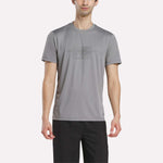 A man wears the Reebok Men's Reebok x F45 Training T-Shirt in Grey 3, featuring performance design and "RS TRAINING" print on the front, paired with black pants and standing against a plain white background.