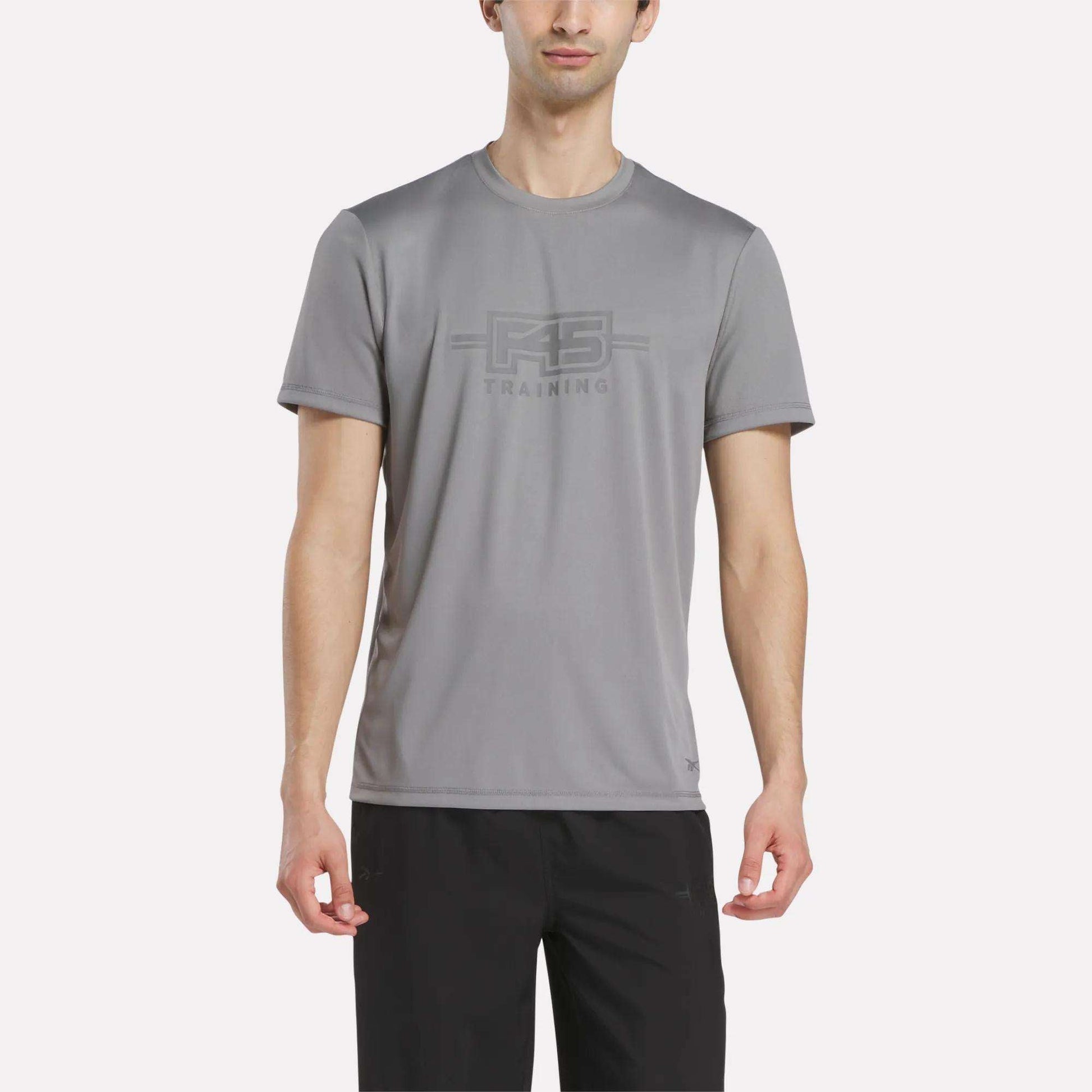 A man wears the Reebok Men's Reebok x F45 Training T-Shirt in Grey 3, featuring performance design and "RS TRAINING" print on the front, paired with black pants and standing against a plain white background.