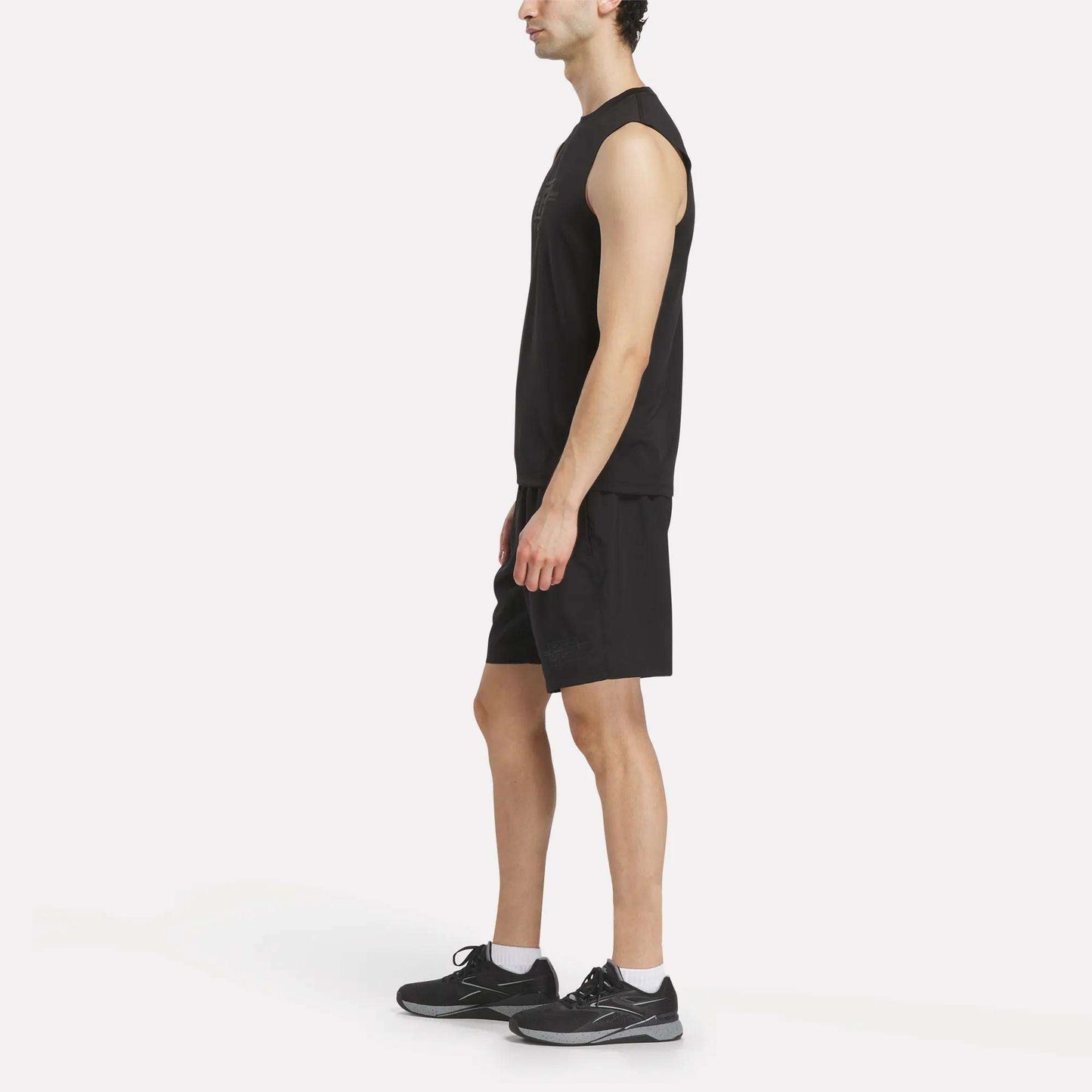 A man stands in profile view against a plain white background, wearing the Reebok x F45 Training Tank Top for men—perfect for high-intensity workouts or an F45 Training session. He pairs it with black shorts and black athletic shoes with white soles.