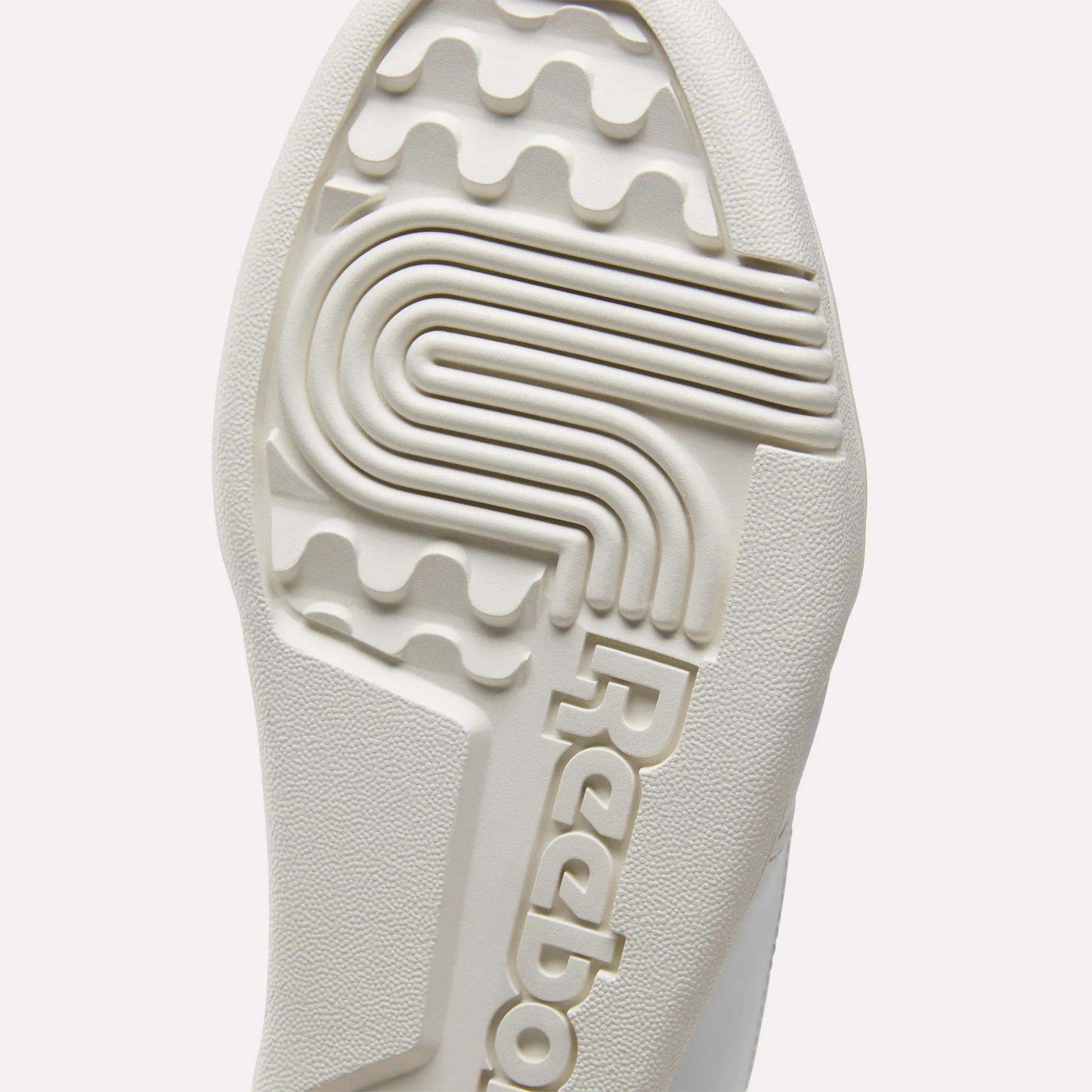 Close-up of the textured sole of white Reebok Workout Plus Shoes for big kids, featuring the brand name and a unique pattern with grooves and ridges for sporty traction.