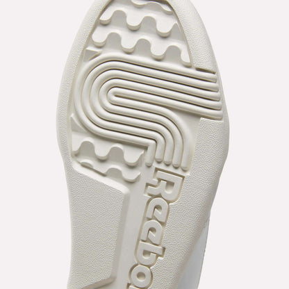 Close-up of the textured sole of white Reebok Workout Plus Shoes for big kids, featuring the brand name and a unique pattern with grooves and ridges for sporty traction.