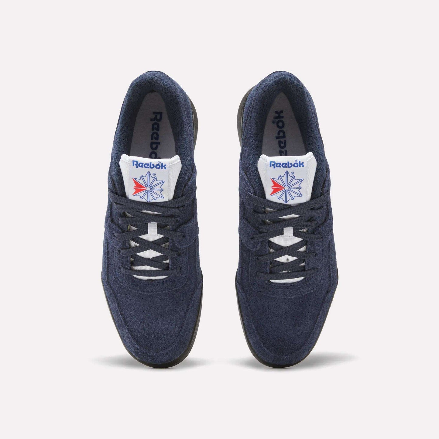 A pair of Reebok Workout Plus Shoes in navy blue with white laces and soles, featuring the Reebok logo and a red-blue star on the white tongue, capture classic '80s style against a light background.