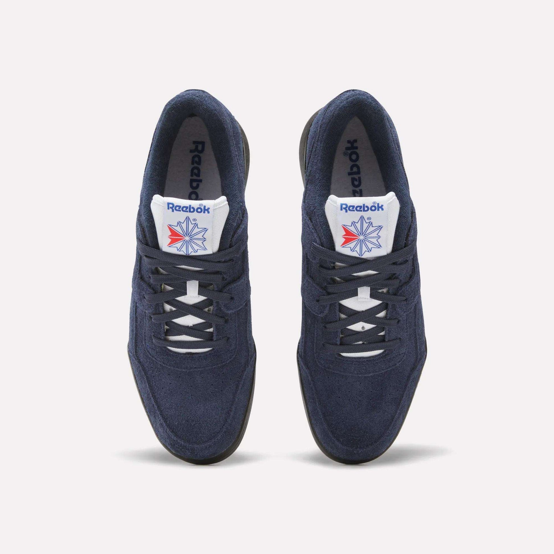 A pair of Reebok Workout Plus Shoes in navy blue with white laces and soles, featuring the Reebok logo and a red-blue star on the white tongue, capture classic '80s style against a light background.