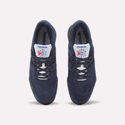 A pair of Reebok Workout Plus Shoes in navy blue with white laces and soles, featuring the Reebok logo and a red-blue star on the white tongue, capture classic '80s style against a light background.