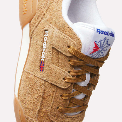 A close-up of tan suede Reebok Workout Plus Shoes with brown laces, a white tongue showing the Reebok logo and Union Jack, and a white and gum sole. Classic '80s fitness style is highlighted with visible stitching and textured suede.