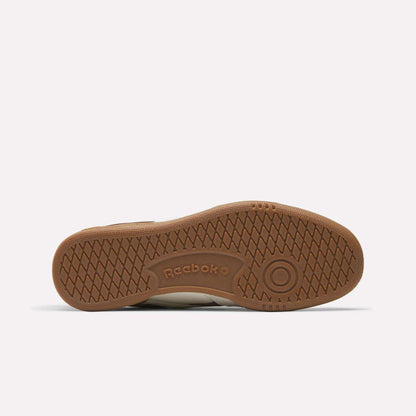 The brown rubber sole of the Reebok Men's Club C Revenge Vintage Shoes is pictured from below, showcasing herringbone tread, circular grips, and a central embossed Reebok logo—a vintage-inspired design.