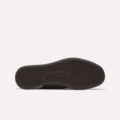 The image shows the sole of a dark Men's Club C 85 Shoe by Reebok, featuring a textured crisscross pattern, a circular toe design, and the embossed Reebok logo at the center.