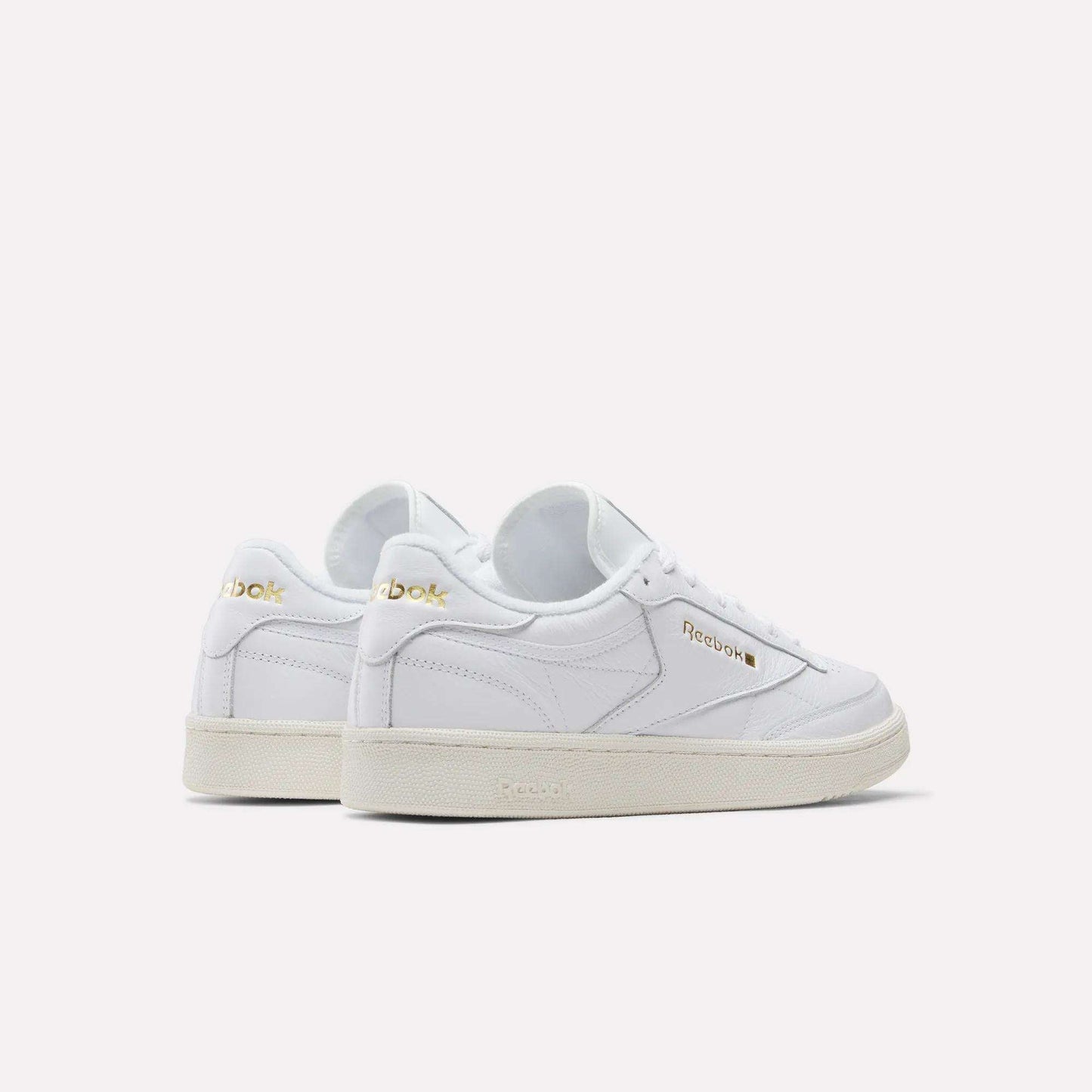 A pair of Reebok Men's Club C 85 Shoes in white garment leather with gold logo accents, white laces, and beige rubber soles—displayed angled to show side and back views against a plain light background.