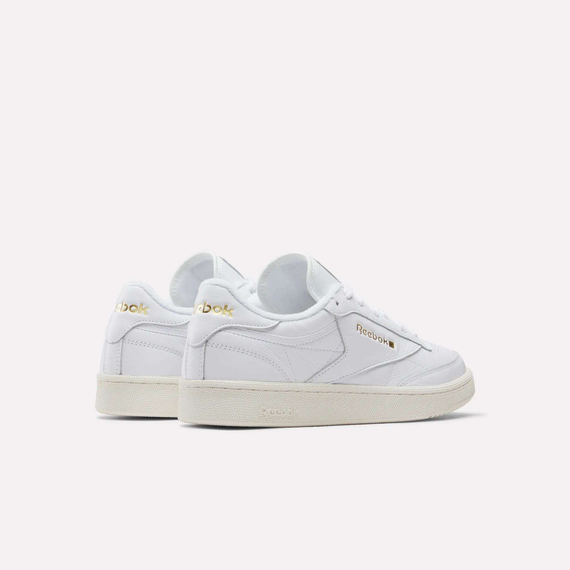 A pair of Reebok Men's Club C 85 Shoes in white garment leather with gold logo accents, white laces, and beige rubber soles—displayed angled to show side and back views against a plain light background.