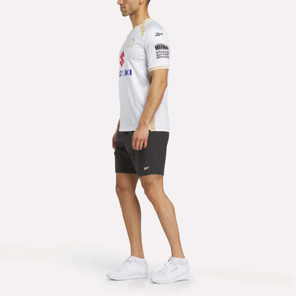 A man stands sideways against a white background wearing the Reebok Men's MK Dons Home Jersey, paired with black shorts and white sneakers.