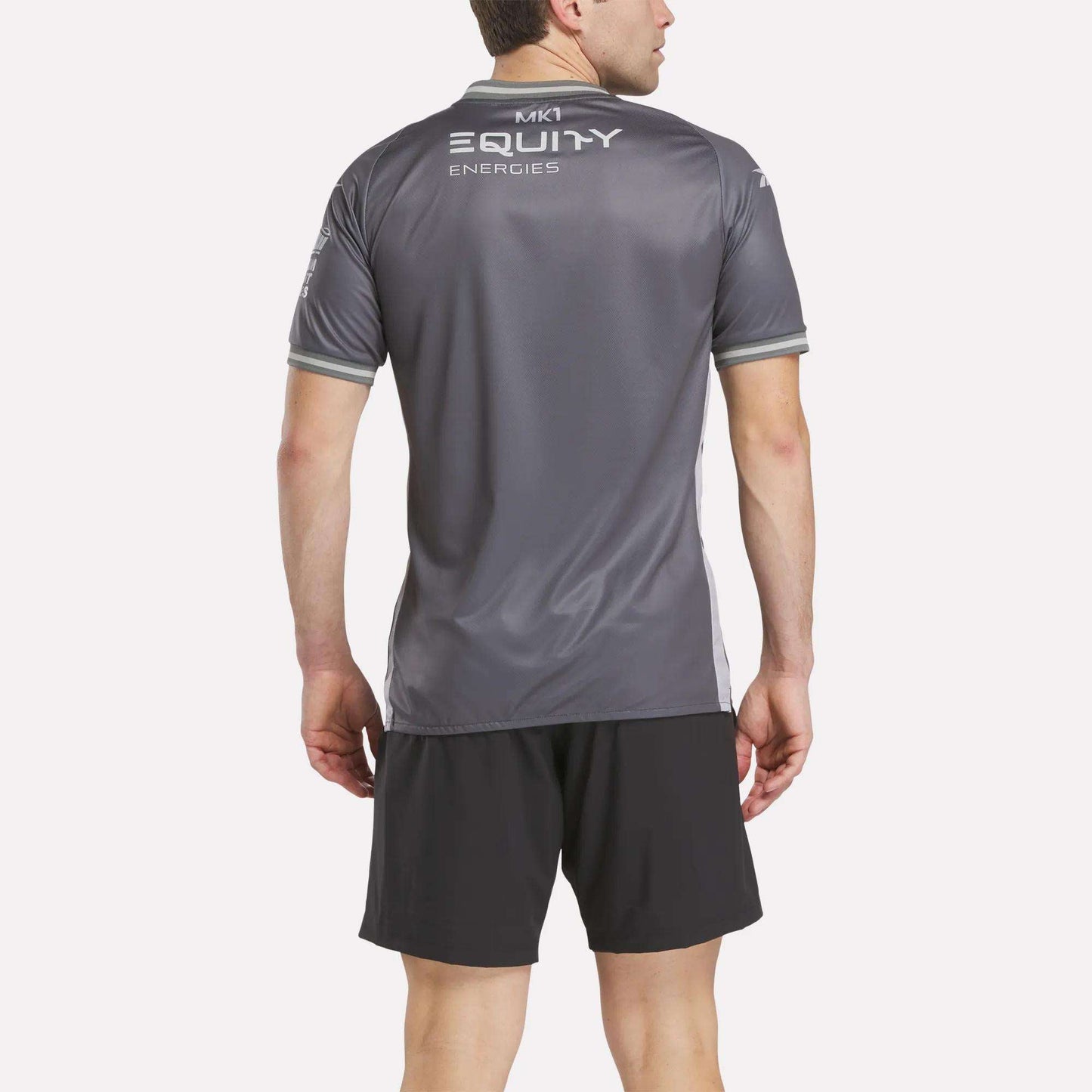 A man faces away, wearing a gray Reebok Men's MK Dons Away Jersey with "MK1 EQUITY ENERGIES" on the back and black athletic shorts, set against a plain light background.