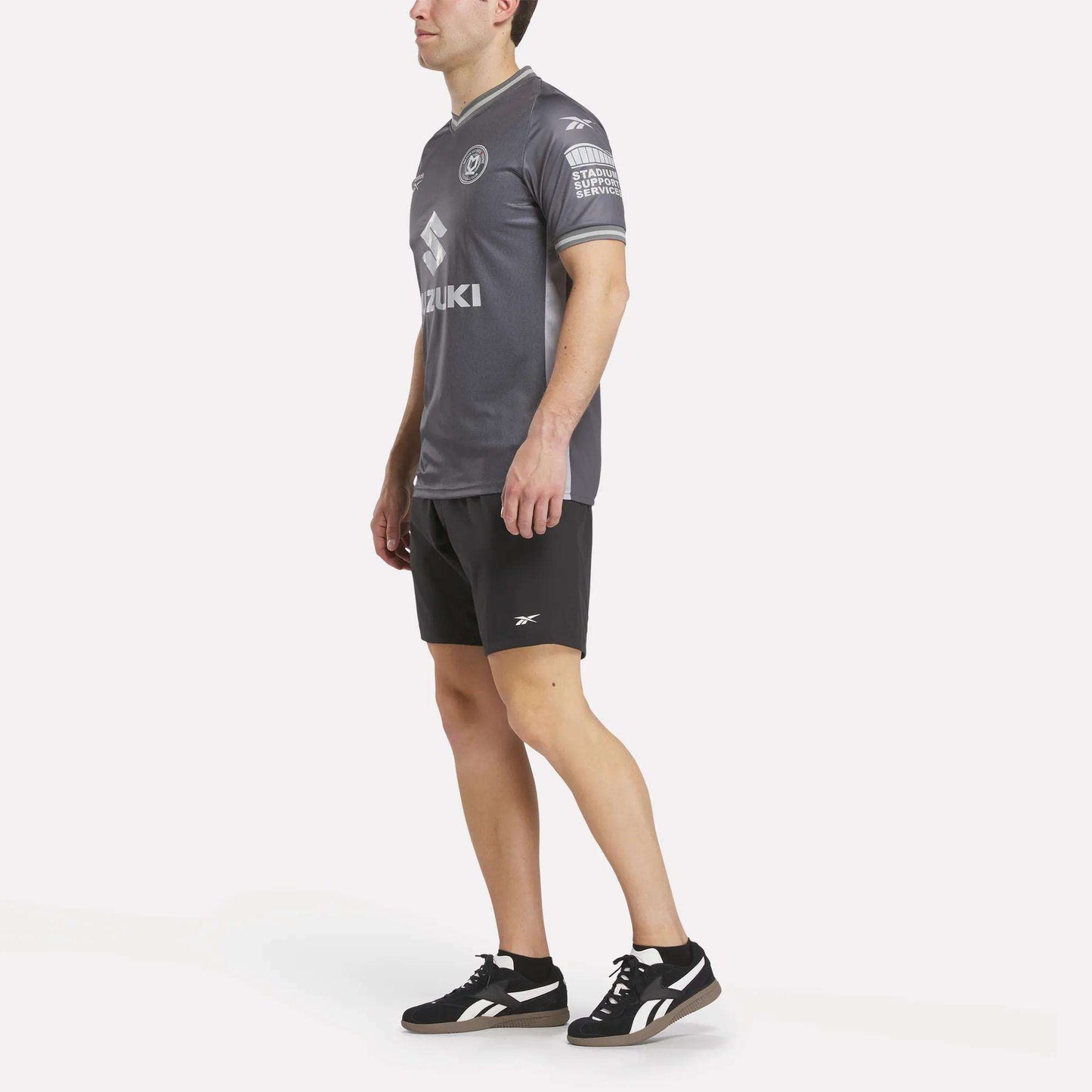 A man wearing a Men's MK Dons Away Jersey, black Reebok shorts, and black Reebok sneakers stands against a plain white background, facing slightly to the side with his arms relaxed by his sides.