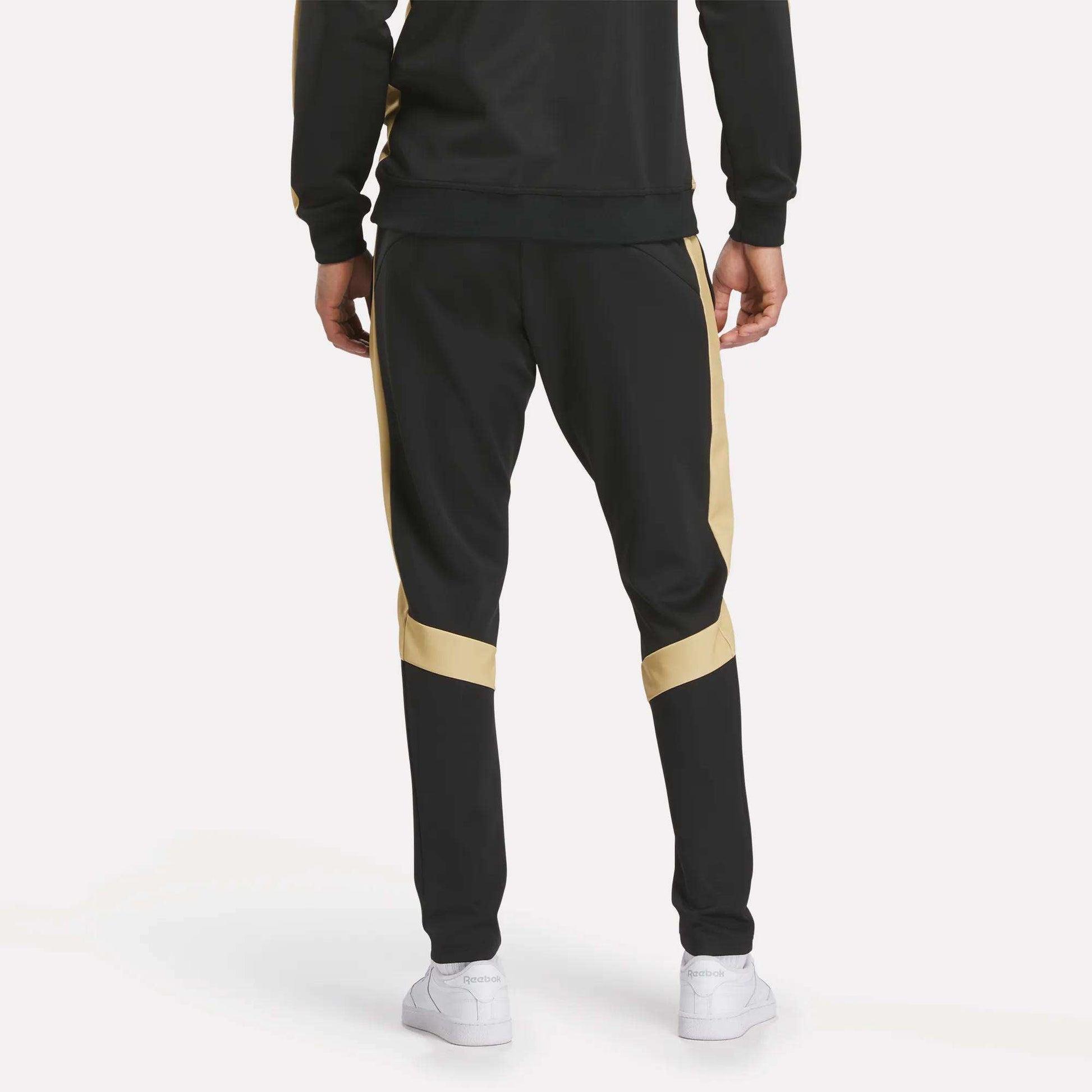 A person is shown from the back against a plain light background wearing Reebok Men's MK Dons Travel Pants with gold side stripes and white sneakers.