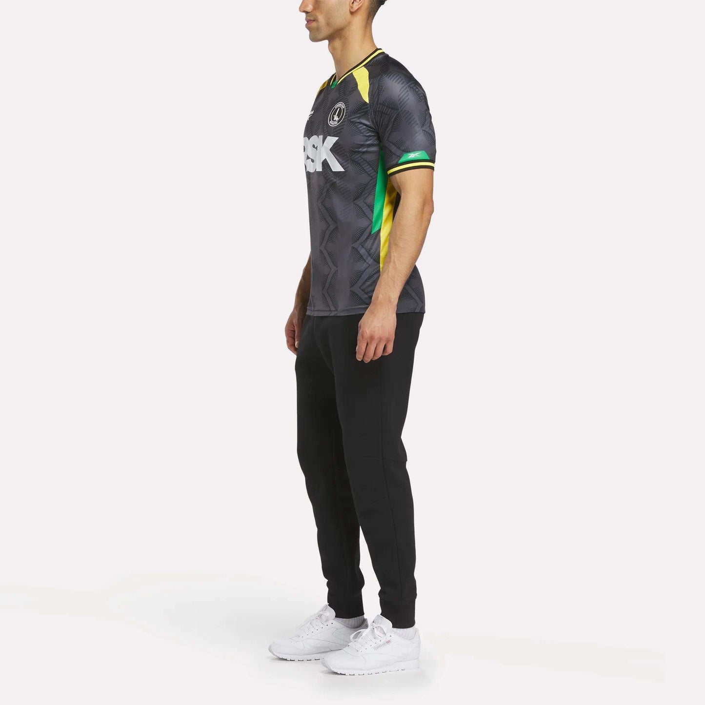 A man stands in profile against a white background, wearing the Reebok Men's Charlton Third Jersey with black pants and white sneakers. The shirt features white lettering plus green and yellow accents inspired by the team's third kit.