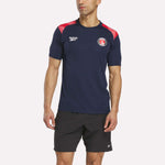 A man wears a navy blue Reebok Men's Charlton Training T-Shirt with red and white shoulder accents, paired with black shorts, standing against a plain light background.