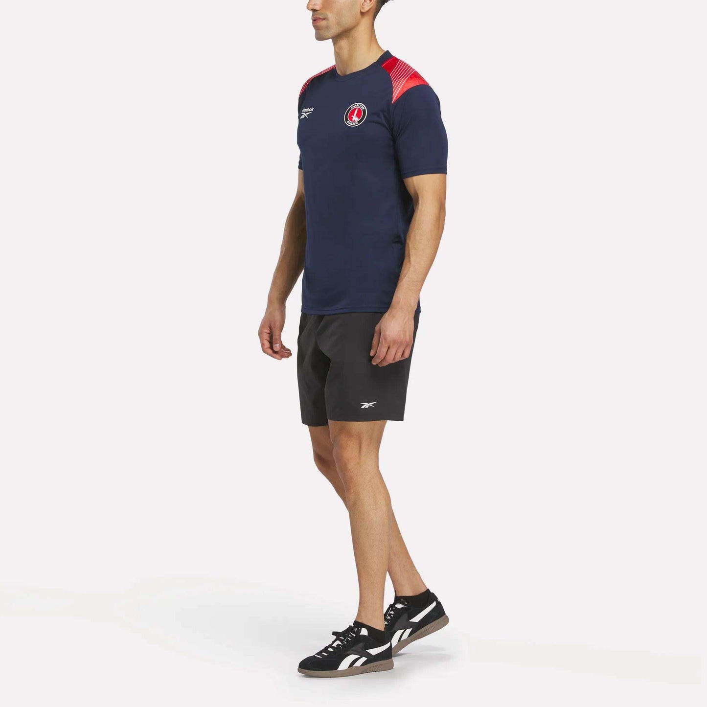 A man faces slightly left, wearing the Reebok Men's Charlton Training T-Shirt in navy blue with red accents, paired with black shorts and black sneakers with white soles. The shirt is made from breathable polyester for comfort.