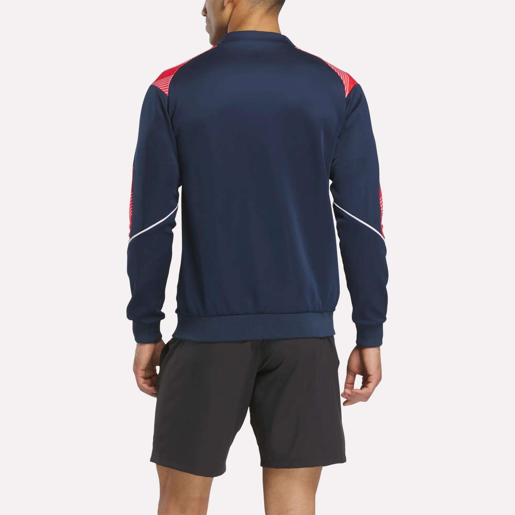 A man stands with his back to the camera in a Reebok Men's Charlton Training Jacket featuring red and white shoulder and sleeve accents, made from polyester doubleknit, paired with black shorts against a plain background.