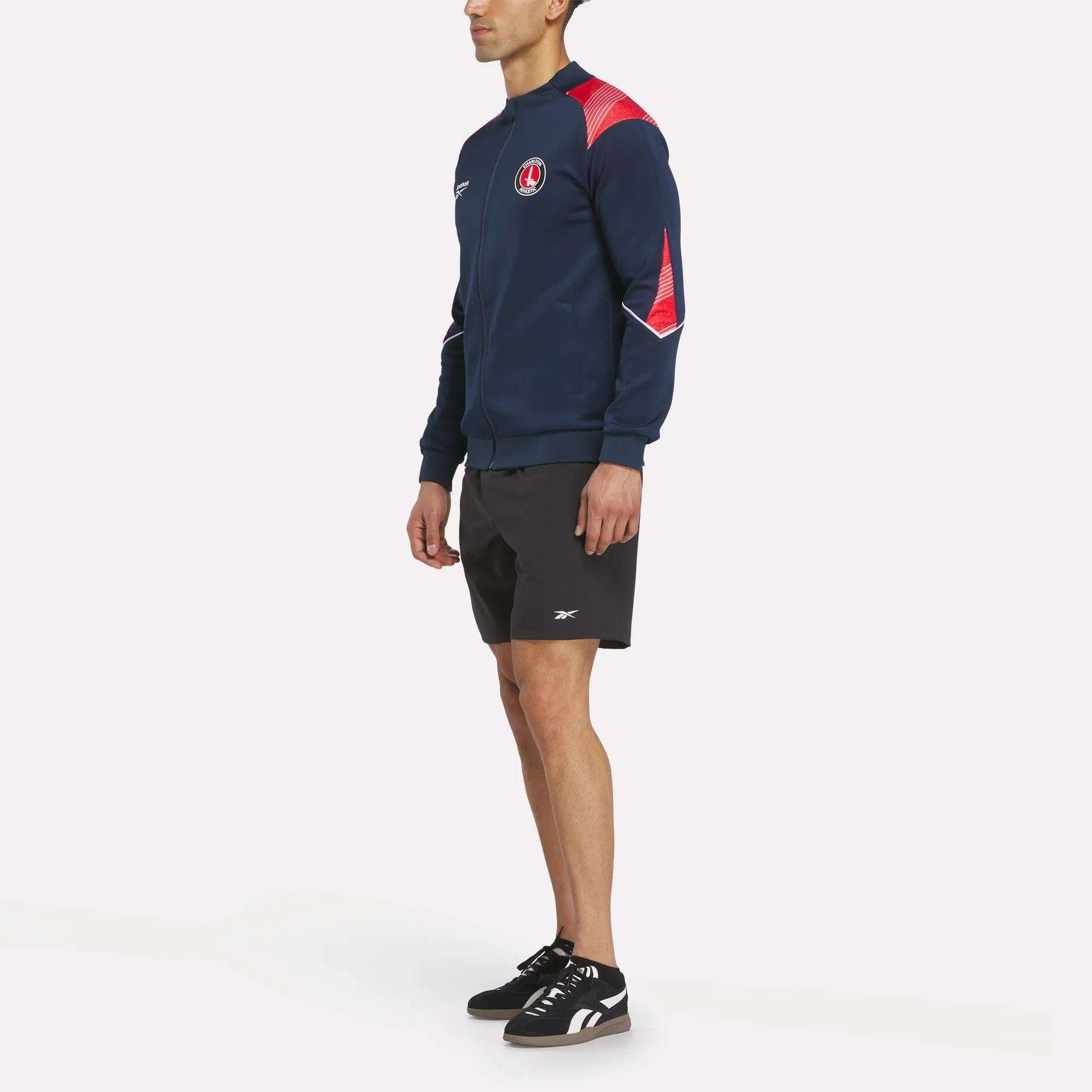 A man in a Reebok Men's Charlton Training Jacket paired with black shorts and sneakers stands facing left against a plain white background.