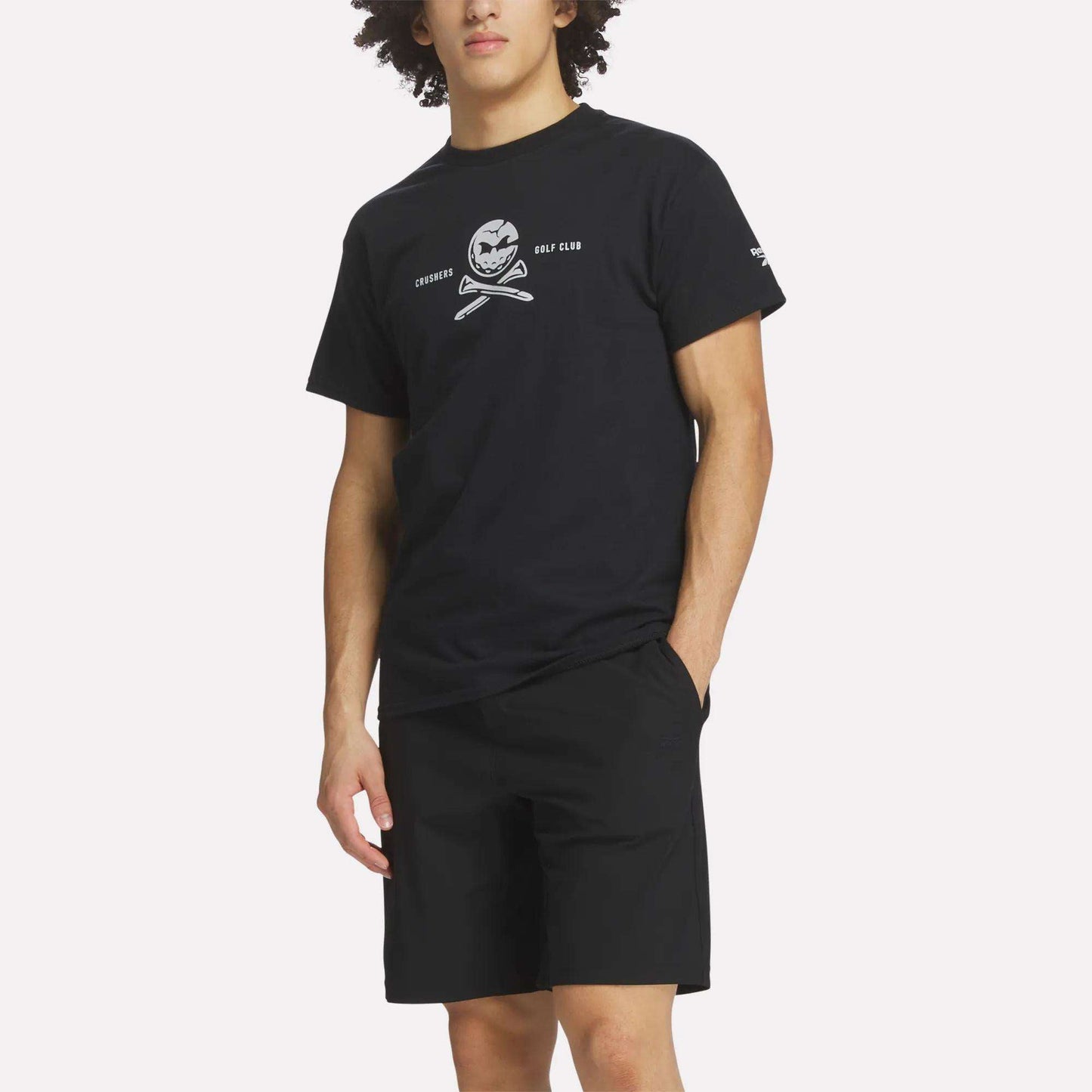 A man wears a Reebok Men's Crushers Tee in black, featuring a skull and crossed golf clubs graphic, paired with black shorts, standing with one hand in his pocket against a plain background.