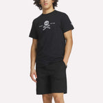 A man wears a Reebok Men's Crushers Tee in black, featuring a skull and crossed golf clubs graphic, paired with black shorts, standing with one hand in his pocket against a plain background.
