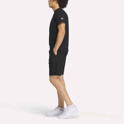 A man stands in profile with hands in pockets, wearing the Reebok Men's Crushers Tee in black, paired with black shorts and white sneakers against a plain white background.