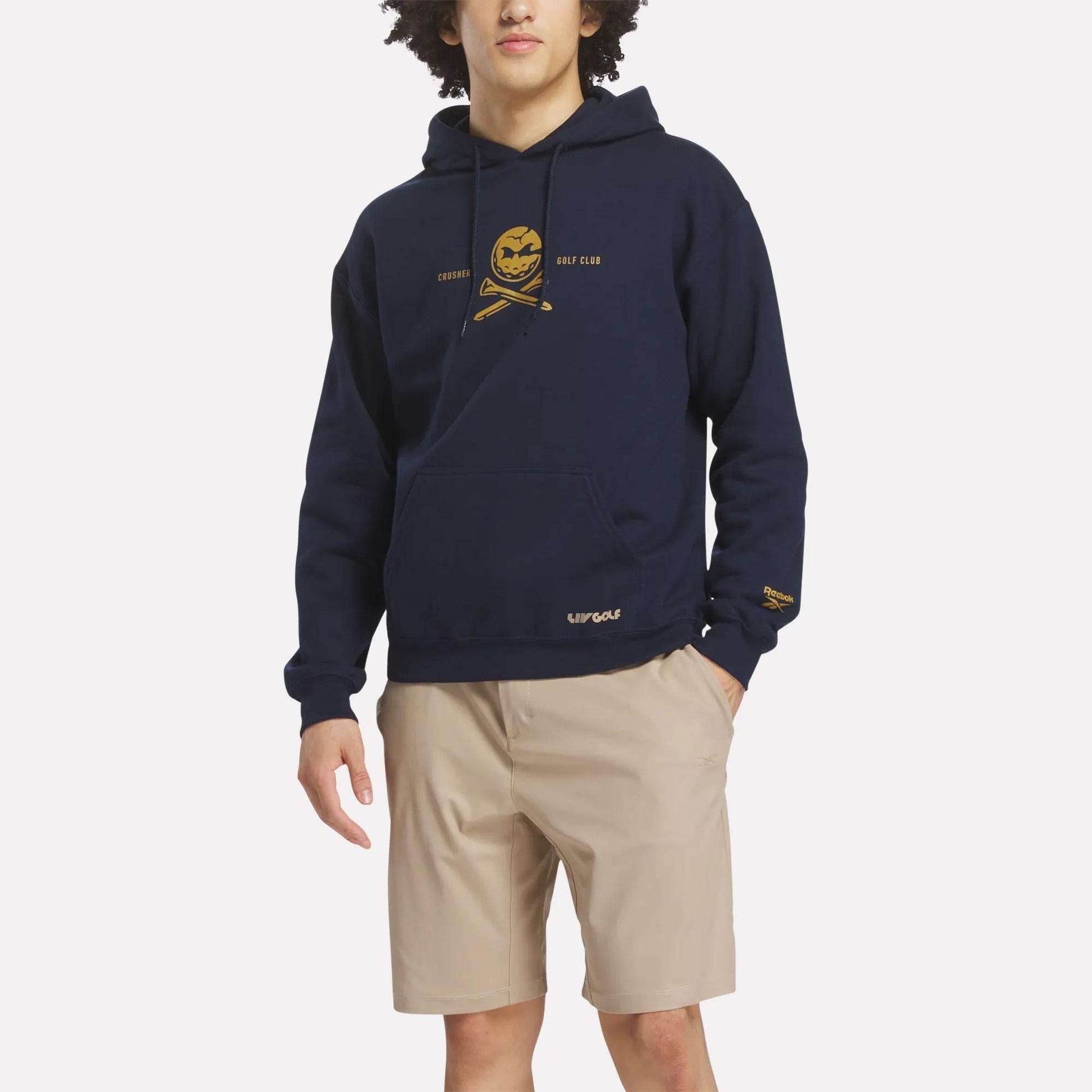 A person wears the Reebok Men's Crushers Hoodie in navy with a yellow chest graphic and text, paired with beige shorts against a plain white background.