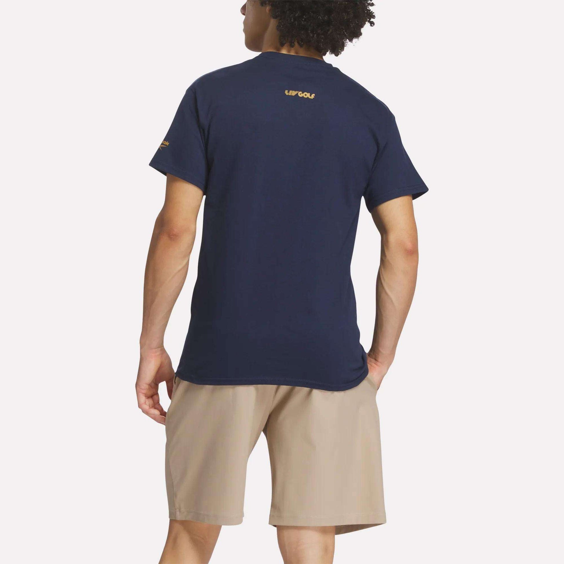 A person faces away against a plain white background, wearing the Reebok Men's Crushers Wordmark Tee in navy blue with yellow text near the collar, paired with tan shorts.